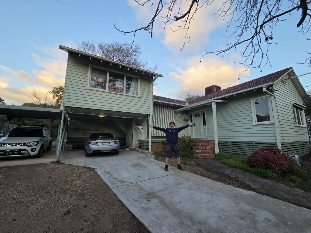Phillip, a white man in his thirties, stands in the driveway of a weatherboard house. He is grinning as he throws his arms out to show off the property.