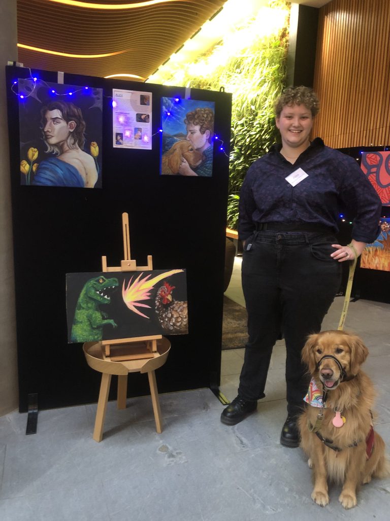 Alex, wearing a dark shirt and black jeans, stands with one hand on their hip in front of their three artworks. Alex is holding the lead of their Golden Retriever dog Daisy