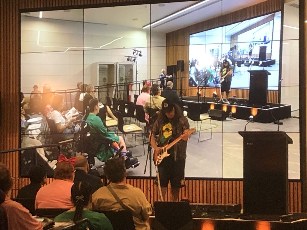 Chris, a man with long hair, plays an electric guitar and sings into a microphone on stage