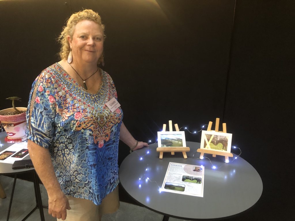 Colette, a woman with blonde hair, stands next to two photographs displayed on small easels on a table