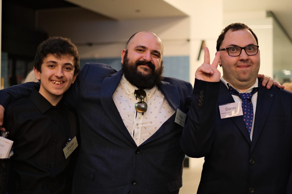 Danni throws a peace sign with his fingers while he stands next to two young men. They are all wearing dark formal wear