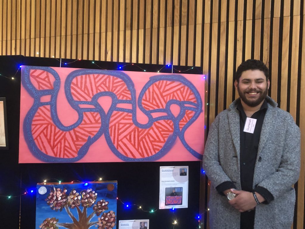 Gurrenah stands smiling next to his large artwork – a pink background with a snake outlined in blue and purple, filled with red strokes
