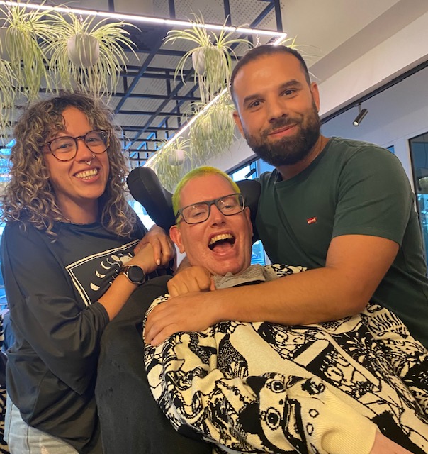 Alan sitting in a chair smiling widely while Stavros hugs him. Gaby is standing behind them smiling