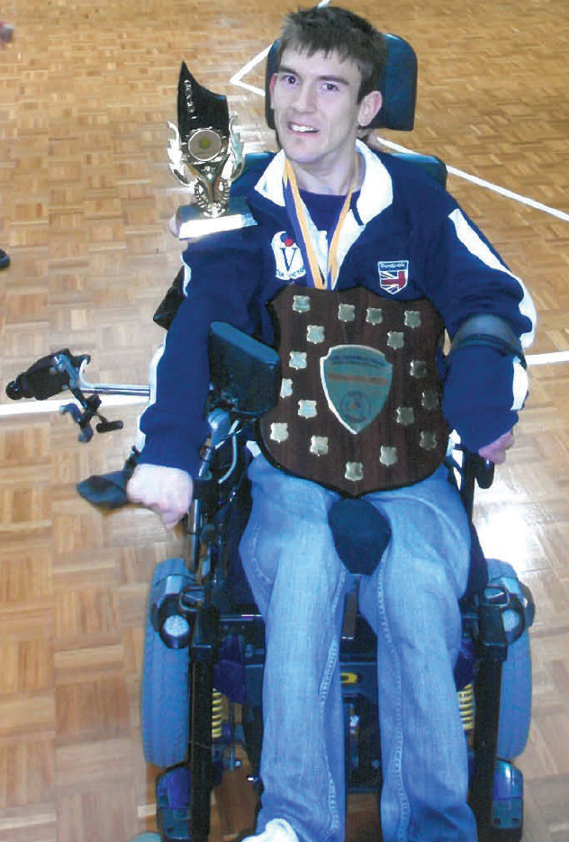 Gavin smiling with his many boccia awards - he has a trophy, a shield and medals