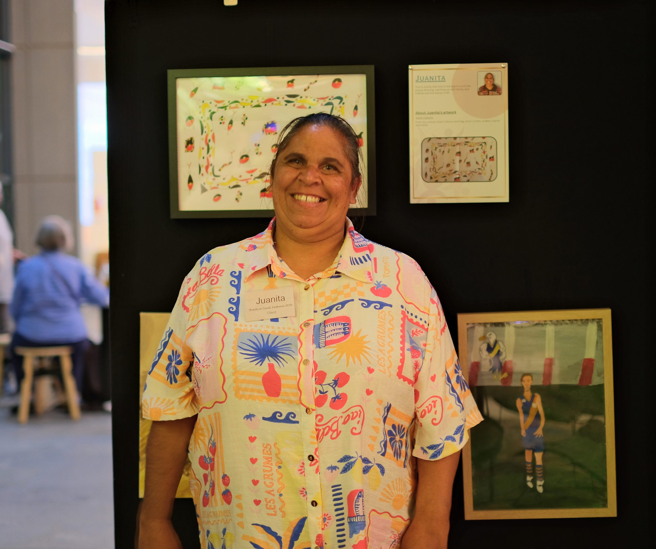 Juanita, a Koori woman, smiles broadly as she stands in front of her artwork which is displayed on a black wall. She is wearing a bright patterned shirt
