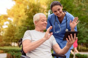 A nurse stands next to an older male patient who is sitting in a wheelchair. They are waving to someone on the man's tablet