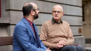 Elderly man in a fawn-coloured jumper and olive shirt sits on a bench talking to a middle-aged man in a blue suit