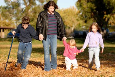 A woman and three young children hold hands while walking through autumn leaves. The child on the left is using a walking stick.